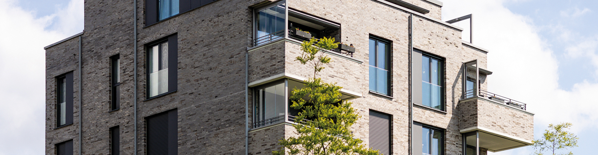 Mehrfamilienhaus mit moderner Architektur und heller Klinkerfassade, großen Fenstern und Balkonen. Ein Baum steht vor dem Gebäude.