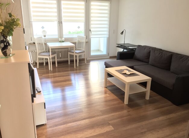 Heller Wohnraum mit Holzboden, dunklem Sofa, Couchtisch und Fernseher. Im Hintergrund ist ein kleiner Essbereich vor großen Fenstern mit viel Tageslicht.