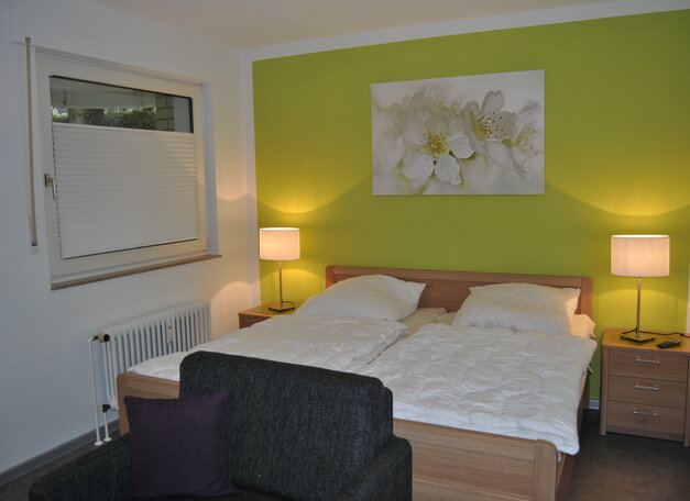Schlafzimmer mit Doppelbett, Nachttischen und zwei Lampen vor einer grünen Akzentwand. Über dem Bett hängt ein Blumenbild, Tageslicht fällt durch ein Fenster.