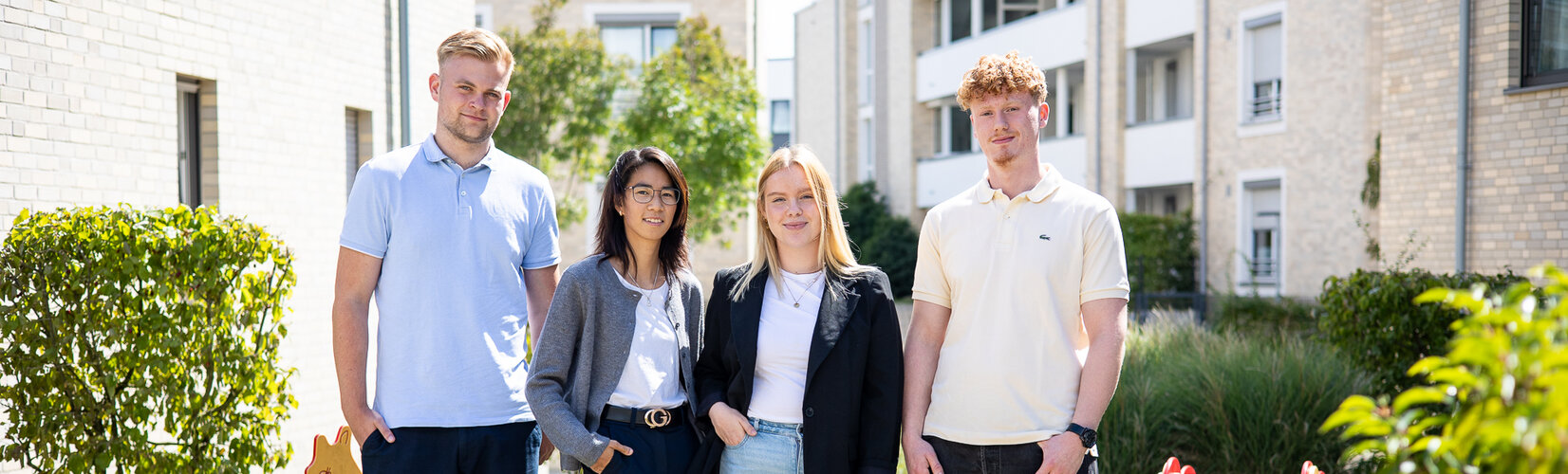 Vier junge Erwachsene stehen im Freien vor Wohngebäuden und Grünflächen und schauen in die Kamera. Sie sind locker gekleidet, die Stimmung wirkt freundlich und offen.