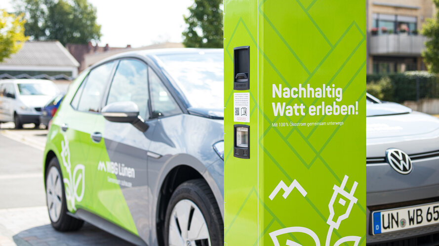 Elektroauto der WBG Lünen steht an einer grünen Ladesäule. Auf der Ladesäule steht der Schriftzug „Nachhaltig Watt erleben!“, auf dem Fahrzeug sind das WBG-Lünen-Logo und ein grünes Design zu sehen.