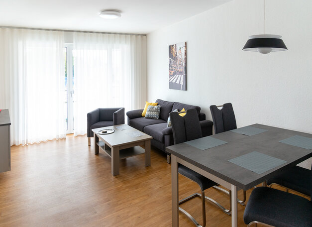 Moderner Wohn- und Essbereich mit dunklem Sofa, Couchtisch und Esstisch mit Stühlen. Große Fenster mit Vorhängen sorgen für viel Tageslicht, der Raum ist hell und freundlich eingerichtet.