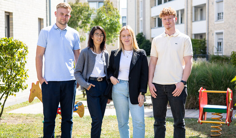 Vier junge Erwachsene stehen im Freien vor Wohngebäuden und Grünflächen und schauen in die Kamera. Sie sind locker gekleidet, die Stimmung wirkt freundlich und offen.