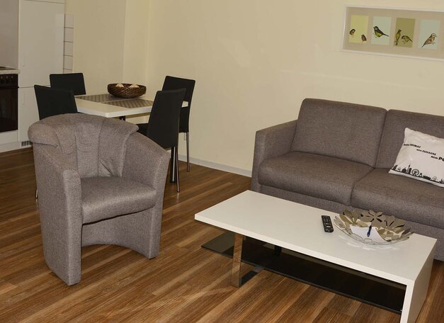 Wohnbereich mit grauem Sofa und Sessel, Couchtisch und Holzboden. Im Hintergrund ist ein kleiner Essbereich mit Tisch und Stühlen sowie eine Küchenzeile zu sehen.