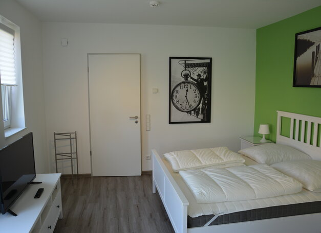 Helles Schlafzimmer mit weißem Bett und Nachttisch, grün gestrichener Akzentwand und Holzfußboden. An der Wand hängen Bilder, links steht ein Fernseher auf einer Kommode.