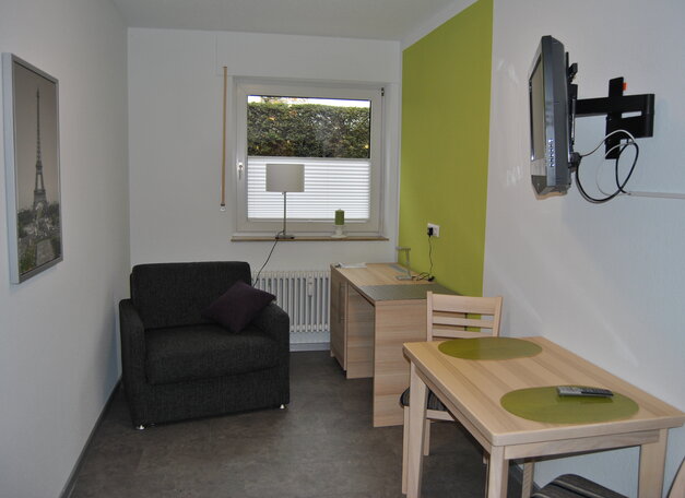 Wohnraum mit Sessel, kleinem Esstisch und Schreibtisch. Eine grüne Akzentwand, ein Fenster mit Tageslicht sowie ein an der Wand montierter Fernseher sind zu sehen.